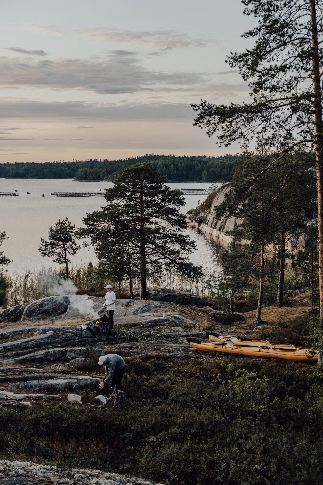 A photograph of the wilderness. There are mountains and a lake in the background. In the foreground are evergreen trees and rock. To the middle left is a person building a fire with smoke going up in the air. This photo is from pexels by alena beliaeva.